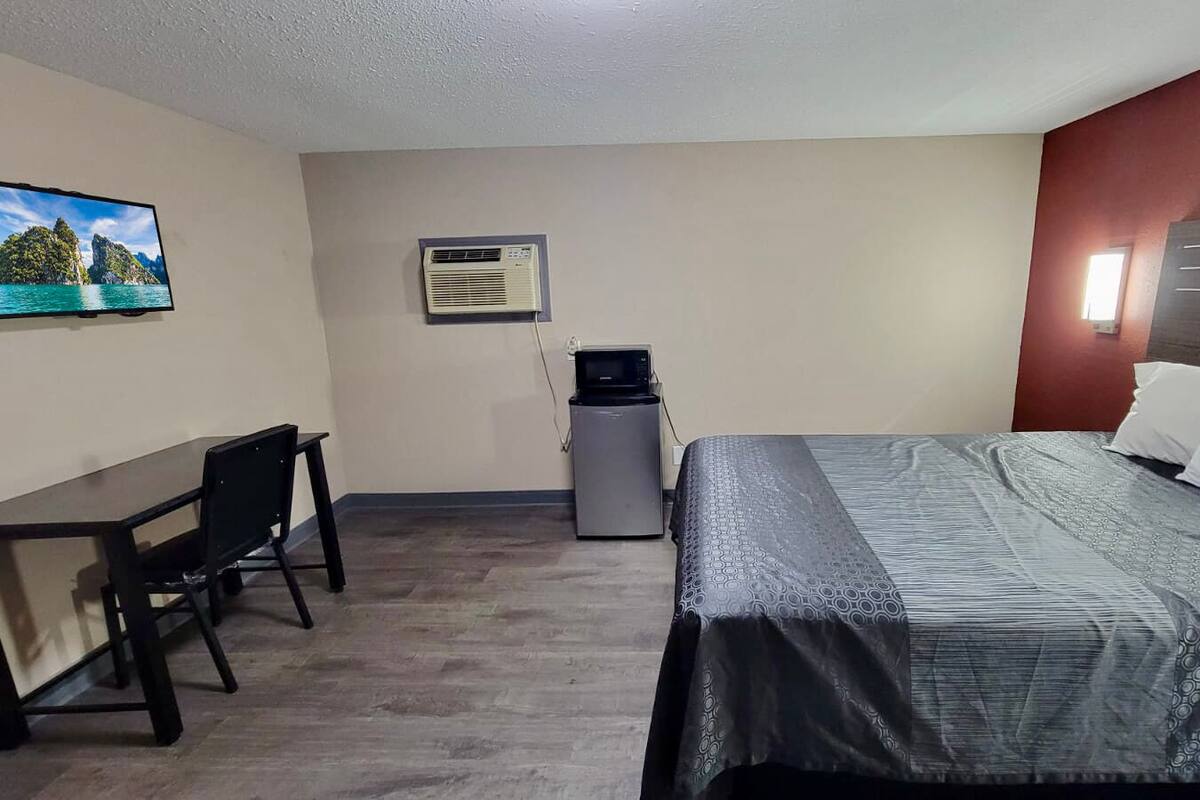 Habitación de hotel con cama, escritorio, silla, equipo de aire acondicionado y televisor en la pared.