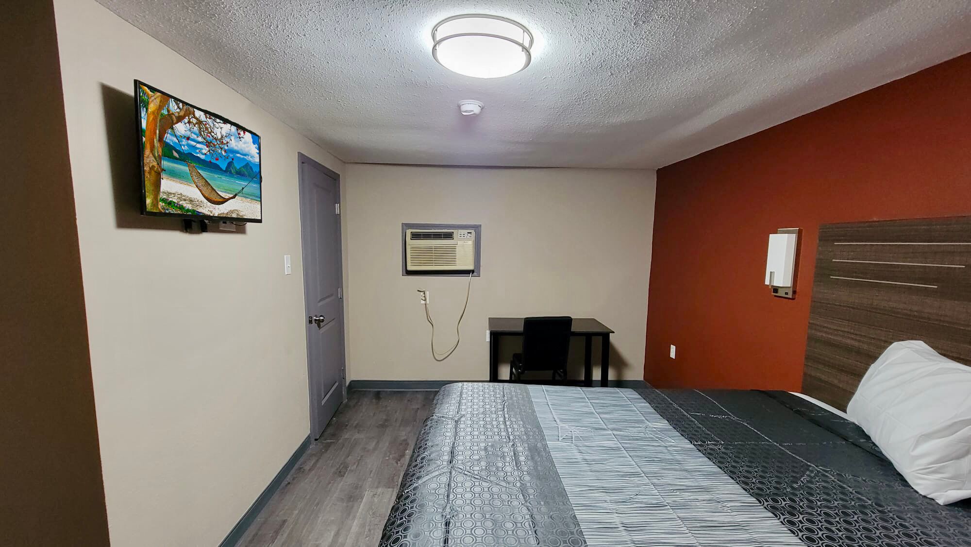 A hotel room with a bed, a desk, an air conditioning unit, and a painting on the wall.
