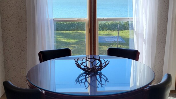 A dining area with a round glass table, six chairs, a chandelier, and a view of a lake through large windows. - Beautiful Lake front property on Big Stone Lake (Clinton)