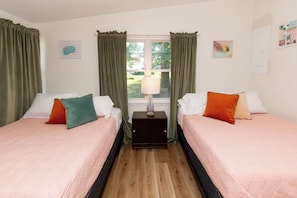 A room with two beds, each with a pillow, a bedside table with a lamp, and a window with curtains. - Mid-Century Modern Themed Surf House (Vermilion)