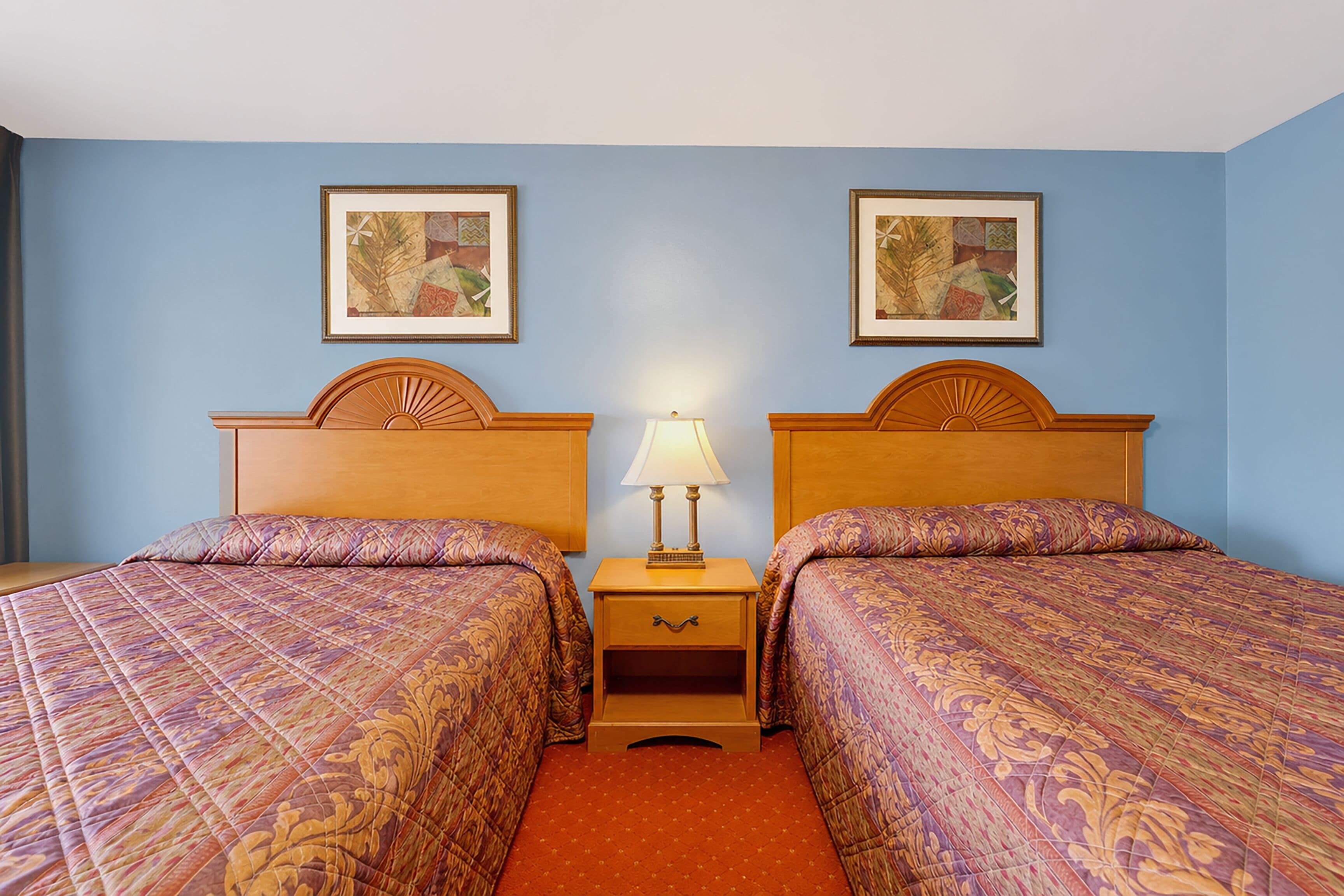 A hotel room with two beds, a nightstand with a lamp, and two framed pictures on the wall.