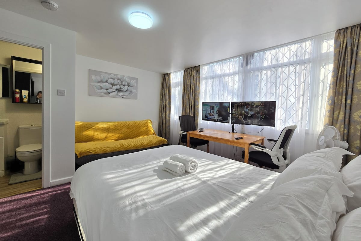 Ein Hotelzimmer mit einem Bett, einem Schreibtisch mit zwei Monitoren, einem Stuhl, einem Fenster mit Vorhängen und einem durchgegangenen Badezimmer.