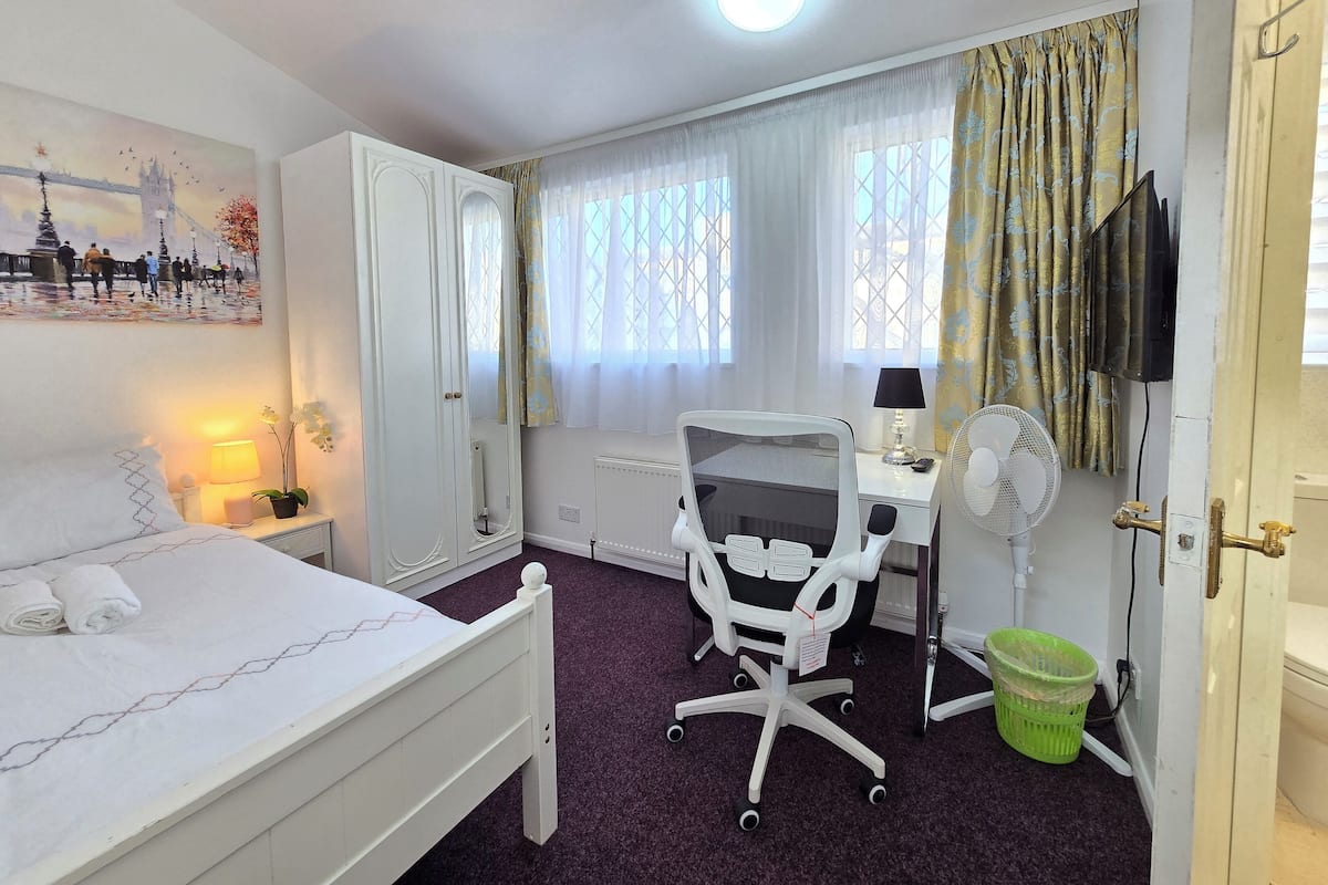 Ein Hotelzimmer mit Bett, Schreibtisch, Stuhl, Standventilator und einem Fenster mit Vorhängen.