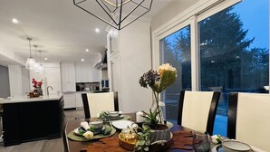 Un área de comedor moderna con una lámpara colgante geométrica, un ventanal con vistas, y una mesa de comedor bien dispuesta con platos, vasos y una pieza central.