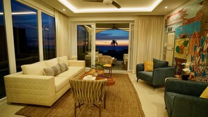 Un salon moderne avec un canapé, des fauteuils et une table basse. On peut admirer le coucher de soleil à travers les portes vitrées.