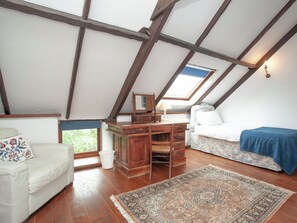 Ein gemütliches Schlafzimmer unter dem Dach mit schrägen Wänden, Holzbalken, einem Schreibtisch mit Lampe, einem Bett mit blauer Decke und einem gemusterten Teppich.
