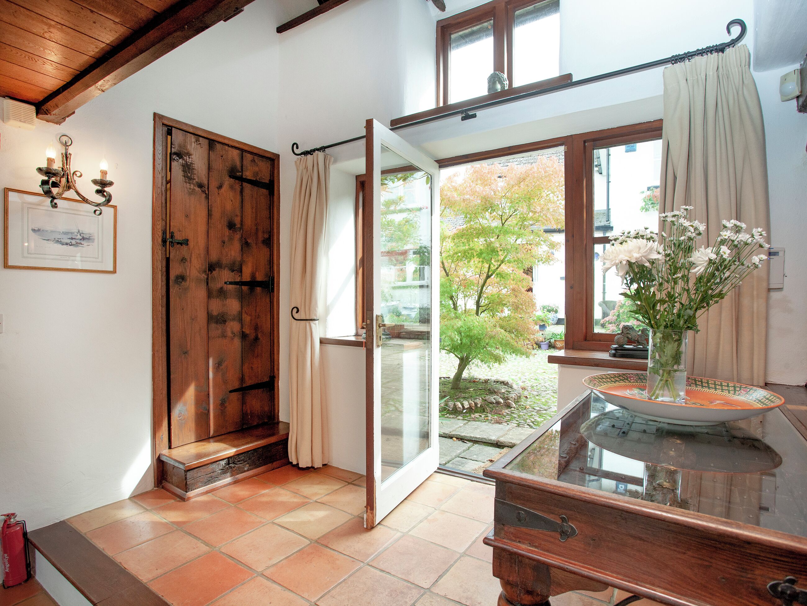 Une pièce avec une porte en bois, une porte vitrée donnant sur l’extérieur, une petite table sur laquelle est posé un vase de fleurs et un tableau encadré au mur.