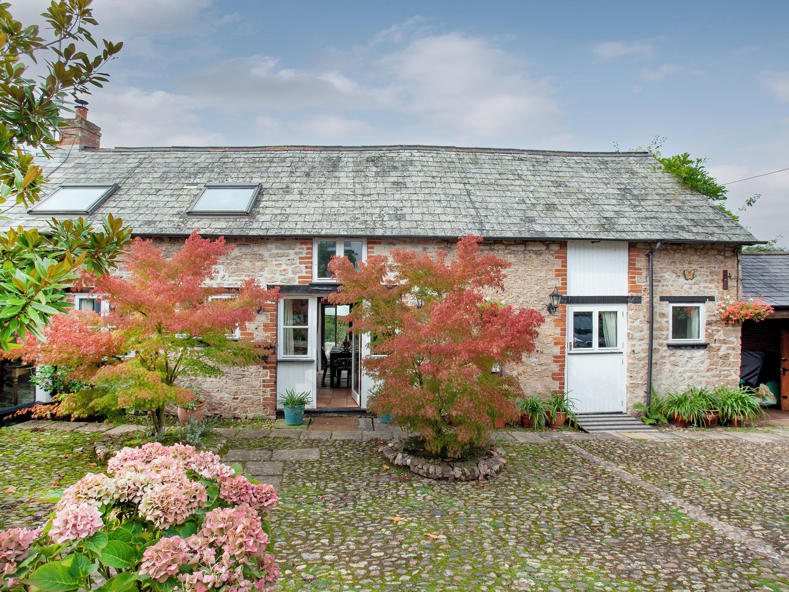 A stone house with a tiled roof, a front door, and a small garden with pink hydrangeas.