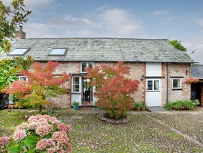 A stone house with a tiled roof, a front door, and a small garden with pink hydrangeas.