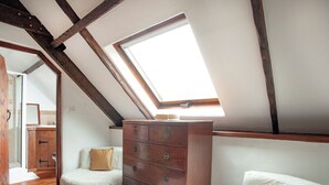 Una habitación con techo inclinado, vigas de madera, un aparador, dos sillas blancas y un tragaluz.