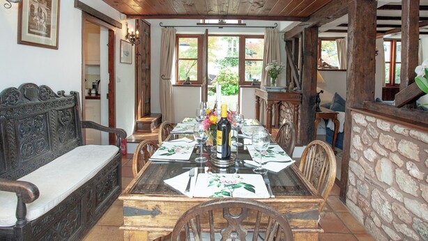 Un comedor con una mesa de madera dispuesta para cuatro personas, rodeada de sillas. Un banco está apoyado contra la pared, y se ve una escalera al fondo.