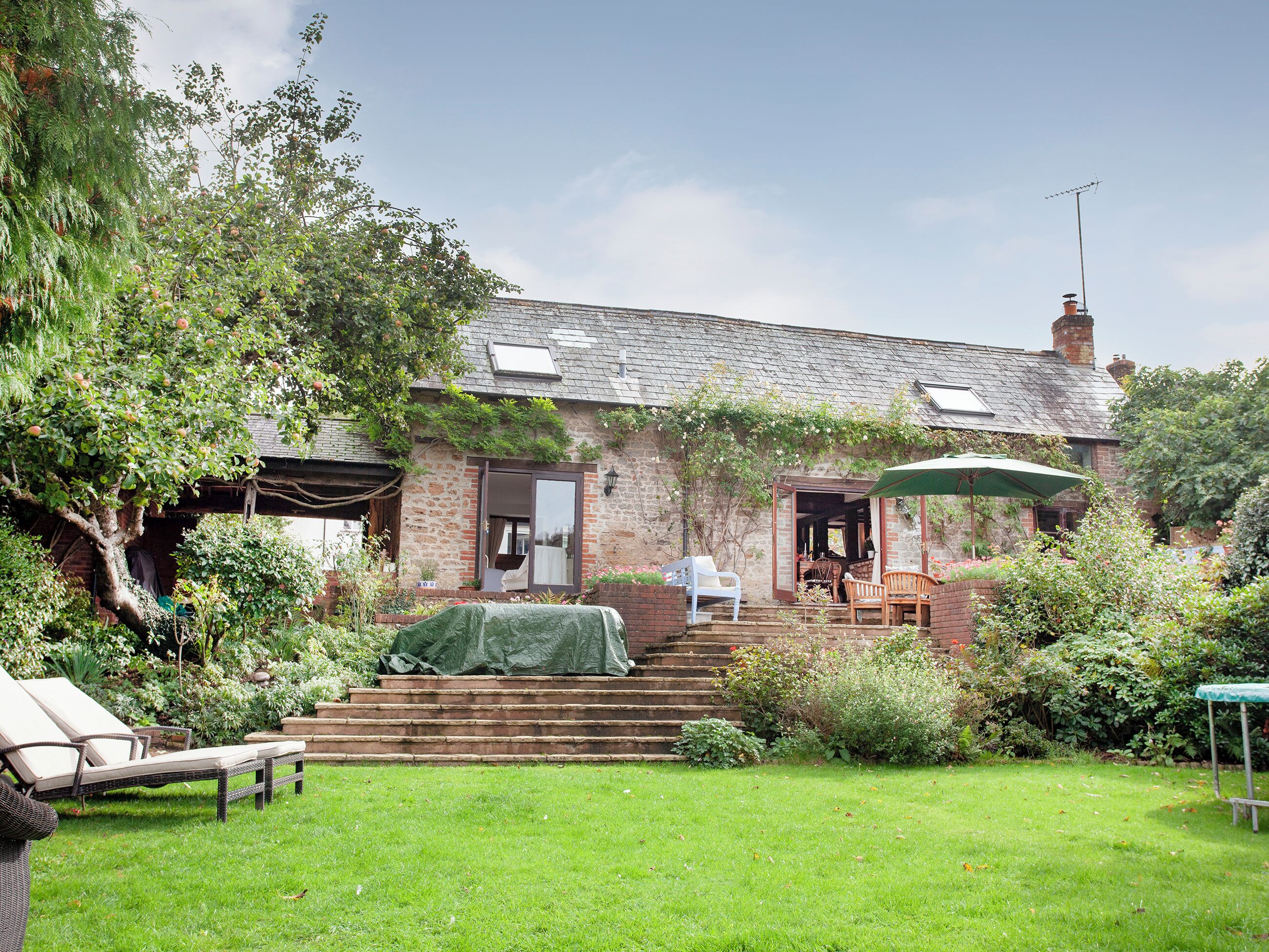 A rustic house with a brick exterior, a covered porch, and a small garden with a lawn and steps leading to the entrance.