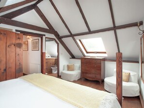 Quarto com teto inclinado, vigas de madeira, claraboia, cama, cômoda, cadeira e um banheiro pequeno.