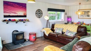 A cozy living room with a brown leather sofa, a small wooden table, a red vintage-style stove, a large wall clock, and a window with a view of greenery. - 2 bedroom accommodation in Hadlow, near Tonbridge (Hadlow, near Tonbridge)