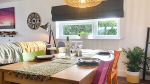 A dining area with a table set for two, a clock, a window with a view of greenery, and a plant in a pot. - 2 bedroom accommodation in Hadlow, near Tonbridge (Hadlow, near Tonbridge)