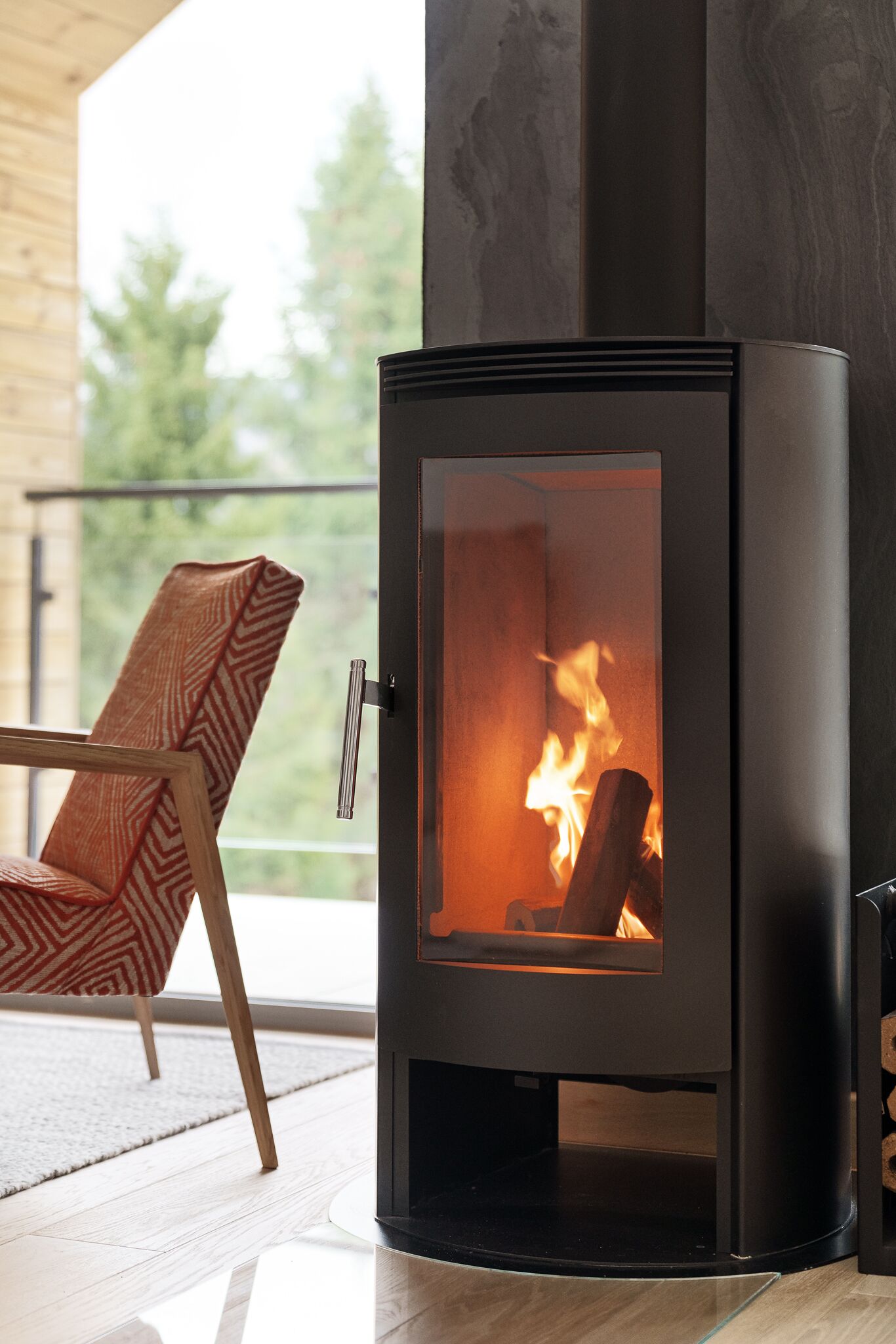 A modern wood-burning stove with a glass door, a wooden chair, and a view of trees through a balcony.