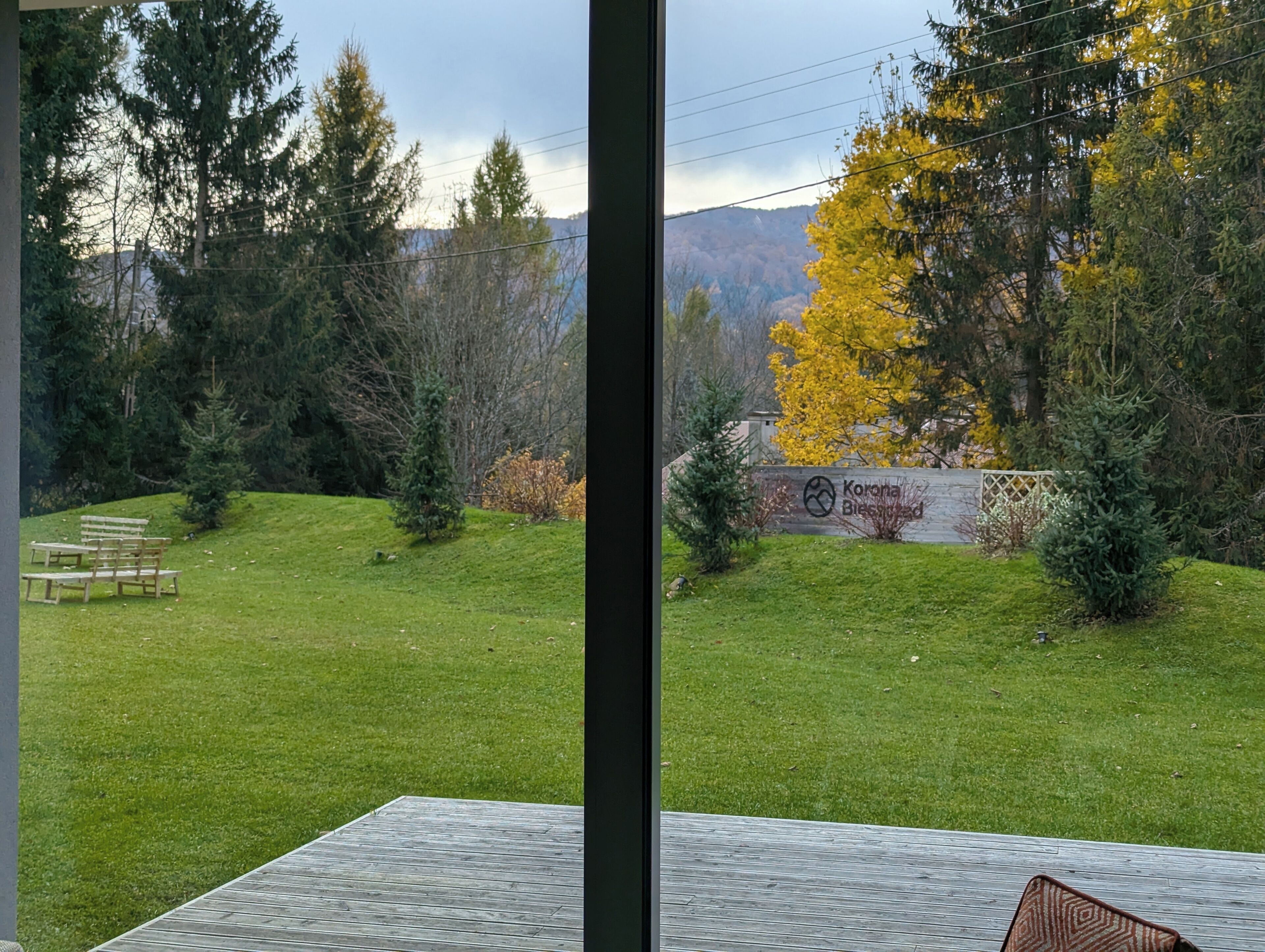 Une vue depuis une fenêtre donnant sur une pelouse bien entretenue, avec un banc en bois, un panneau indiquant « Corona Blissed » et, en arrière-plan, des arbres et des montagnes.