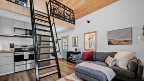 A modern interior with a loft-style kitchen, a staircase, a sofa, and a painting on the wall. - Hygge Up North Bungalow (Bellaire)