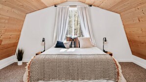A bedroom with a sloped ceiling, a bed with patterned bedding, a window with sheer curtains, and a small bedside table with a lamp. - Hygge Up North Bungalow (Bellaire)