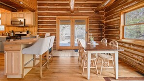 L’intérieur d’une cabane en rondins comprend une table à manger en bois, des chaises, un coin cuisine avec un four à micro-ondes et une grande fenêtre.