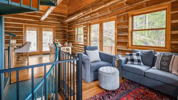 L’intérieur d’une cabane en rondins comprend un coin repas, un salon avec un canapé et un pouf, ainsi qu’un escalier doté d’une rampe.