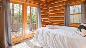 Une chambre située dans un chalet en rondins, avec un grand lit, un balcon avec un fauteuil et une vue sur la forêt.