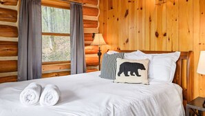 Une chambre dans un chalet en rondins, avec un lit, deux oreillers, une fenêtre avec des rideaux et une lampe.