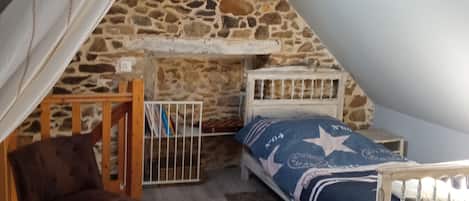 Quarto com teto inclinado, parede de pedra, cama com colcha estampada em formato de estrela, cadeira de madeira e uma prateleira branca pequena.