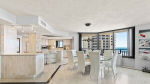 Cuina moderna amb una illa central, zona de dinar amb cadires blanques i un gran finestral que ofereix vistes a edificis i al cel.
