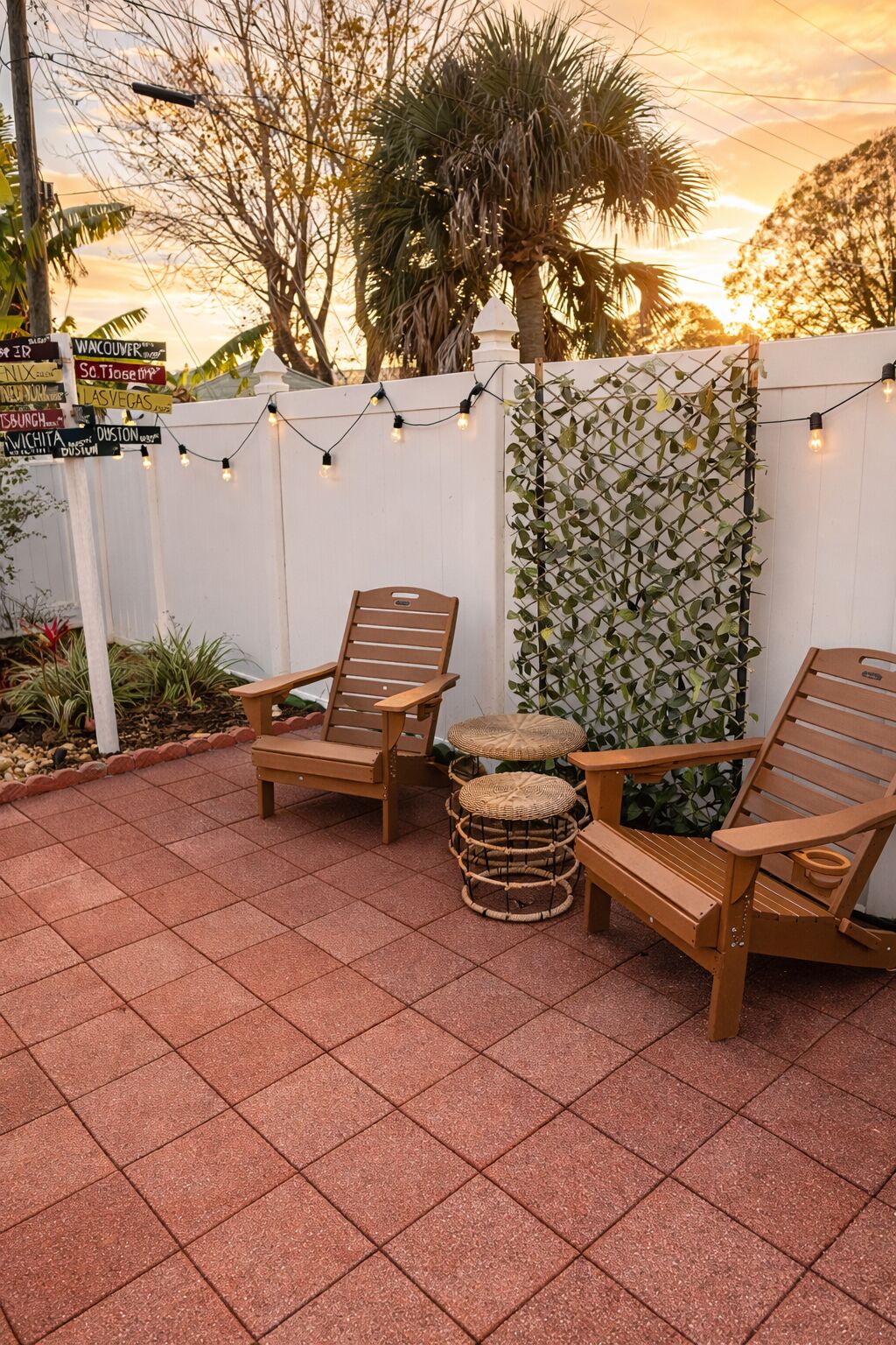 A patio area with wooden chairs and a woven stool, surrounded by a white fence and string lights.