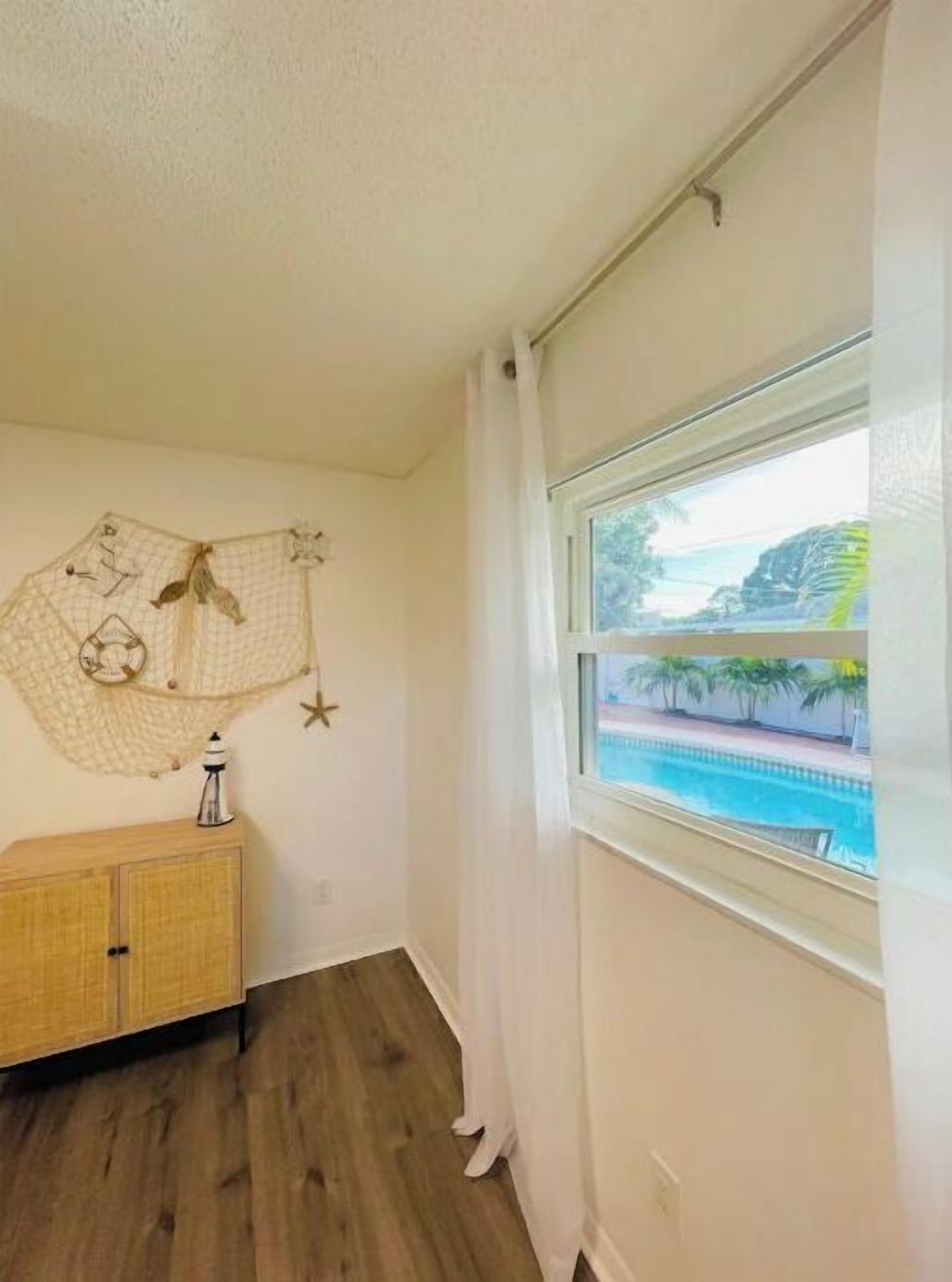 A room with a wooden floor, a white wall with a decorative wall hanging, a wooden cabinet, and a window with a view of a pool and palm trees.