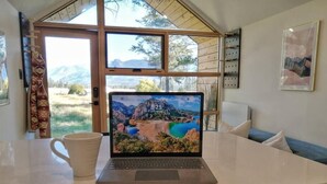 Ein Laptop auf einem Tisch mit einer malerischen Tapete, einer Tasse und Blick auf Berge durch große Fenster.