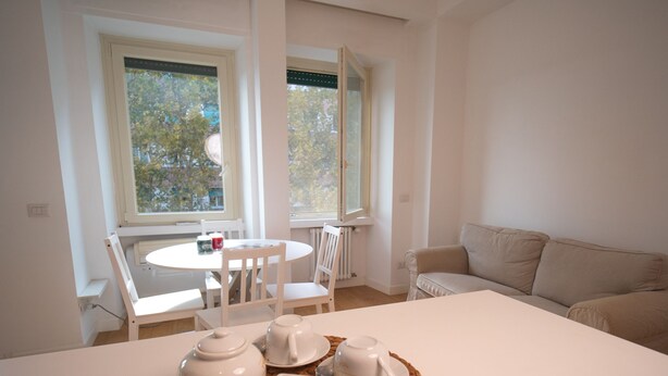 Une pièce moderne et minimaliste, avec une table blanche dressée pour le thé, un canapé beige et une vue sur les arbres à travers de grandes fenêtres.