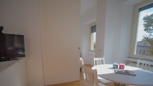 Une pièce lumineuse avec un plancher en bois, des murs blancs, une table à manger avec des chaises, un téléviseur fixé au mur et une fenêtre donnant sur des immeubles.