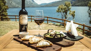 Pullo El Enemigo Cabernet Franc -punaviiniä vuodelta 2017, lasillinen punaviiniä sekä ateria, johon kuuluu empanadoja ja pannulla valmistettu ruoka, kaikki tarjoiltuna puupöydällä järven rannalla, taustalla vuoristo.