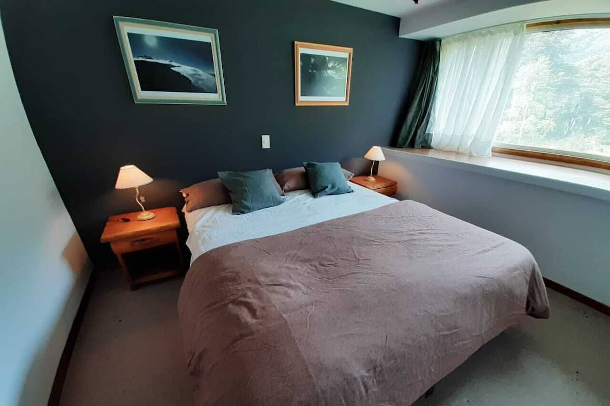 A bedroom with a large bed, two bedside tables with lamps, a window with curtains, and two framed pictures on the wall.