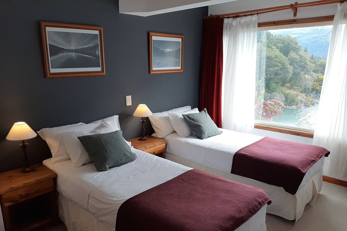 A hotel room with two beds, a nightstand with a lamp, and a window with a view of a mountainous landscape.