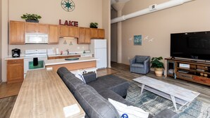 A modern living room with a sectional sofa, a coffee table, a flat-screen TV, and a kitchen area with wooden cabinets and appliances. - Condo with Lake & Cedar Point View (Sandusky)