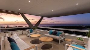 Eine moderne Lounge-Bereich im Freien mit Blick auf den Sonnenuntergang über dem Meer, ausgestattet mit weißen Sofas, runden Holztischen und blauen Kissen.