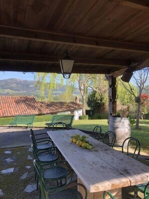 An outdoor covered patio with a wooden table and chairs, a lamp, and a view of a tiled roof and greenery in the background. - Beautiful Stone House, La Llosuca (AS)