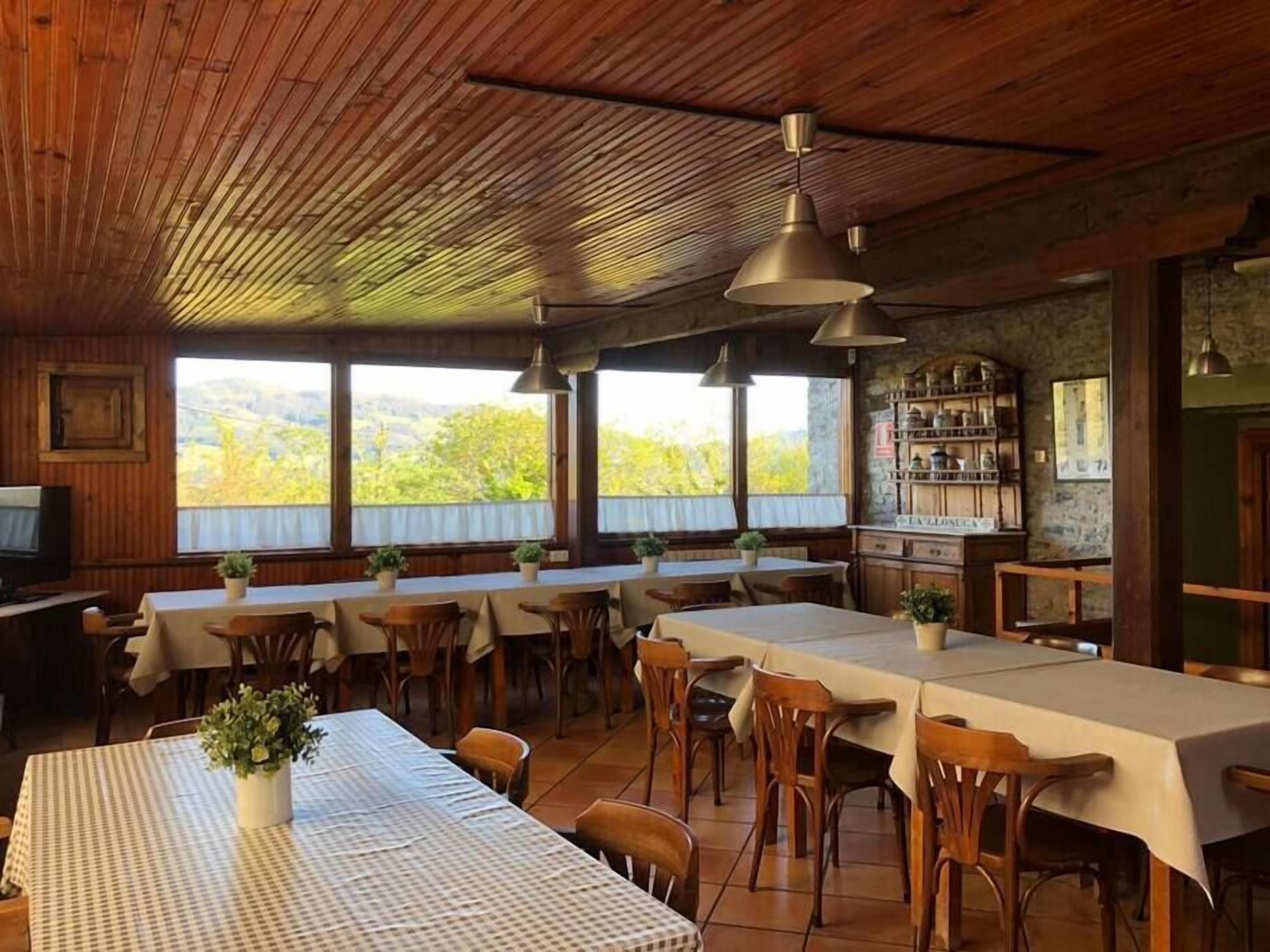 Un comedor rústico con mesas y sillas de madera, ventanas amplias que ofrecen vistas al exterior e iluminación colgante.