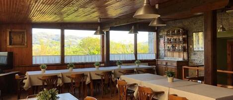 Un comedor rústico con mesas y sillas de madera, ventanas amplias que ofrecen vistas al exterior e iluminación colgante.