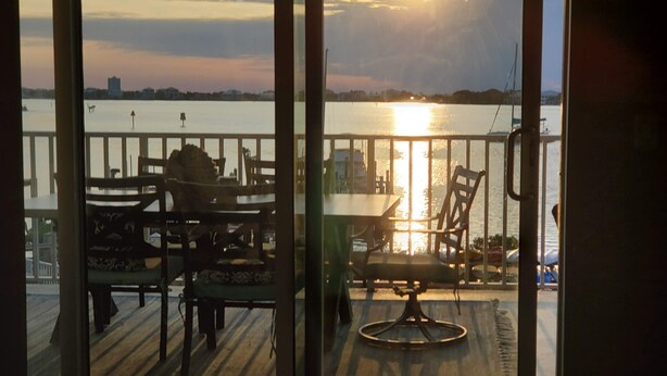 Uitzicht vanuit een kamer met een groot raam op een terras met een tafel, stoelen, een leuning en een waterpartij.