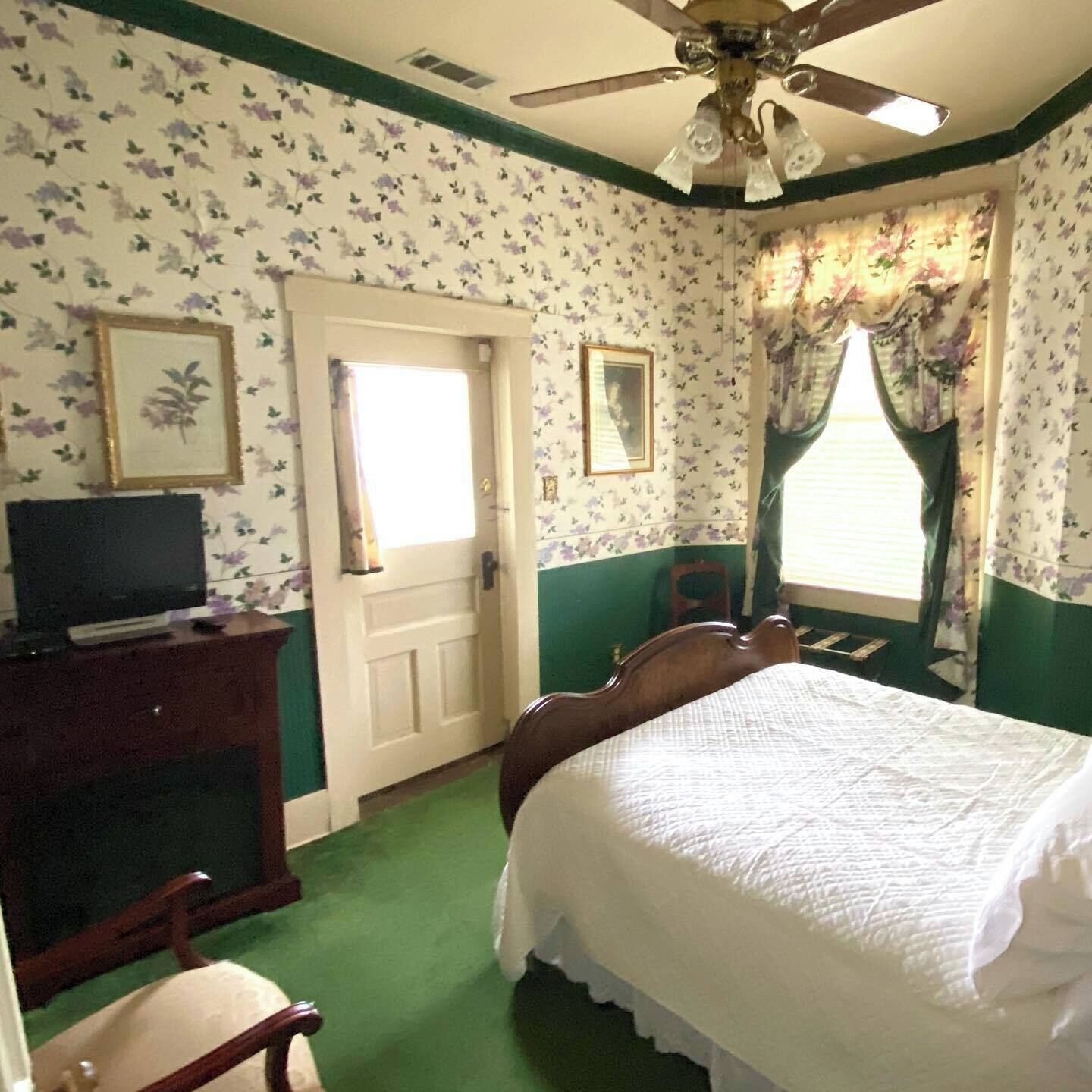 A bedroom with floral wallpaper, a bed, a television on a stand, a ceiling fan, and a window with curtains.