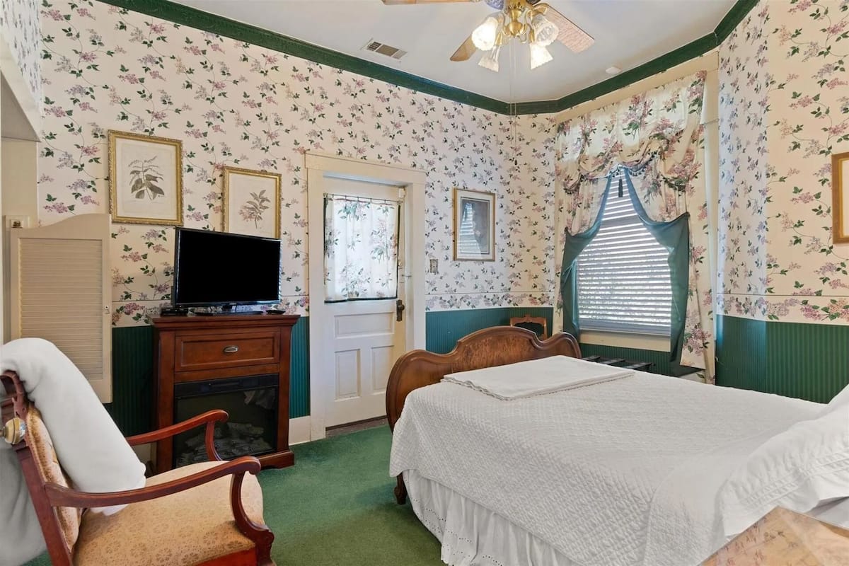 A bedroom with floral wallpaper, a bed, a television on a wooden cabinet, a chair, and a window with curtains.