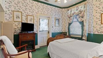 Quarto com papel de parede floral, uma cama, uma televisão sobre um móvel de madeira, uma cadeira e uma janela com cortinas.