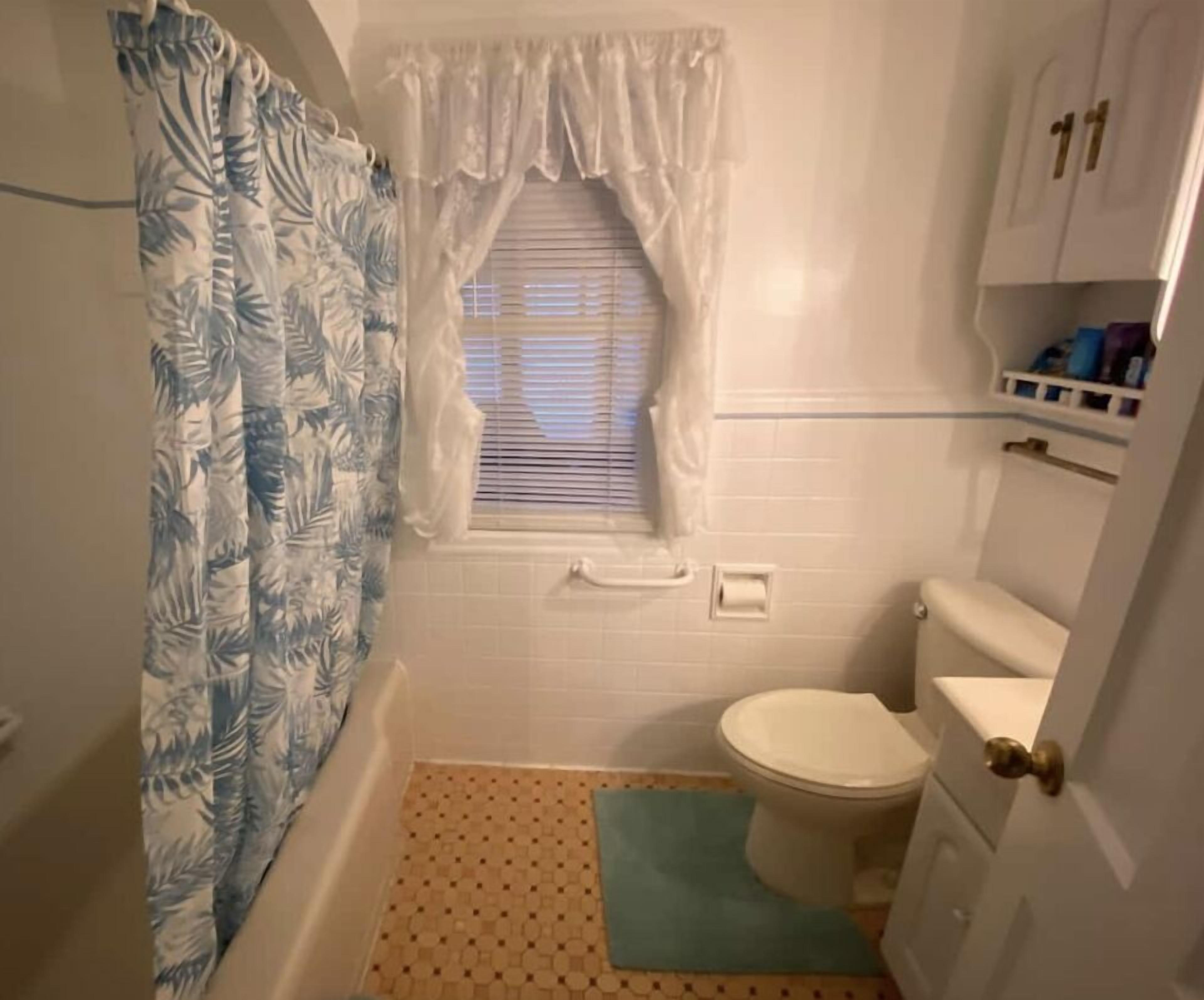 Ein kleines Badezimmer mit Toilette, einem Fenster mit Jalousien, einer Duschvorhang und einem gefliesten Boden.