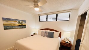 Ein ordentlich eingerichtetes Schlafzimmer mit Bett, Nachttischen, Deckenventilator, einem Fenster mit Jalousien und einem Bild an der Wand.