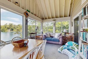 A sunroom with a view of a lake, a dining table with chairs, a sofa, and a bookshelf.
