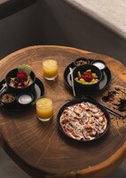 Una mesa de madera con un desayuno que incluye un bol de fruta, un vaso de zumo de naranja, un plato de comida y un cuenco pequeño de yogur.